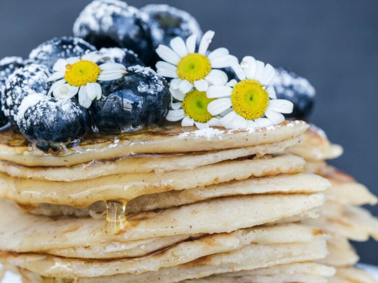 Stack of pancakes topped with blueberries and daisies, drizzled with honey, highlighting a delicious and visually appealing breakfast option.