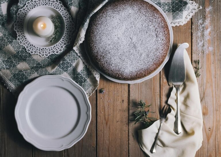 Cake dusted with icing sugar on a white plate, surrounded by a candle, cutlery, and decorative cloth, evoking a warm baking atmosphere related to dessert recipes.