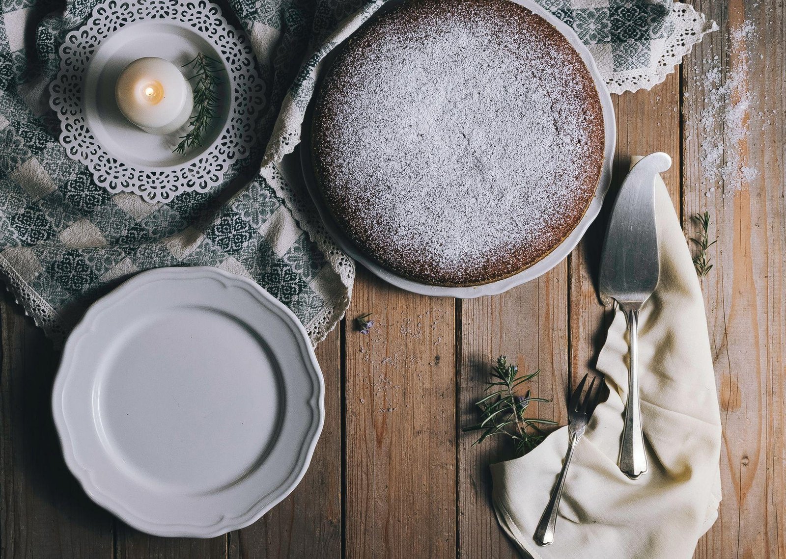 Chocolate spice cake on a decorative plate with powdered sugar, surrounded by a candle, utensils, and a patterned cloth, complementing a blog post about dessert recipes.