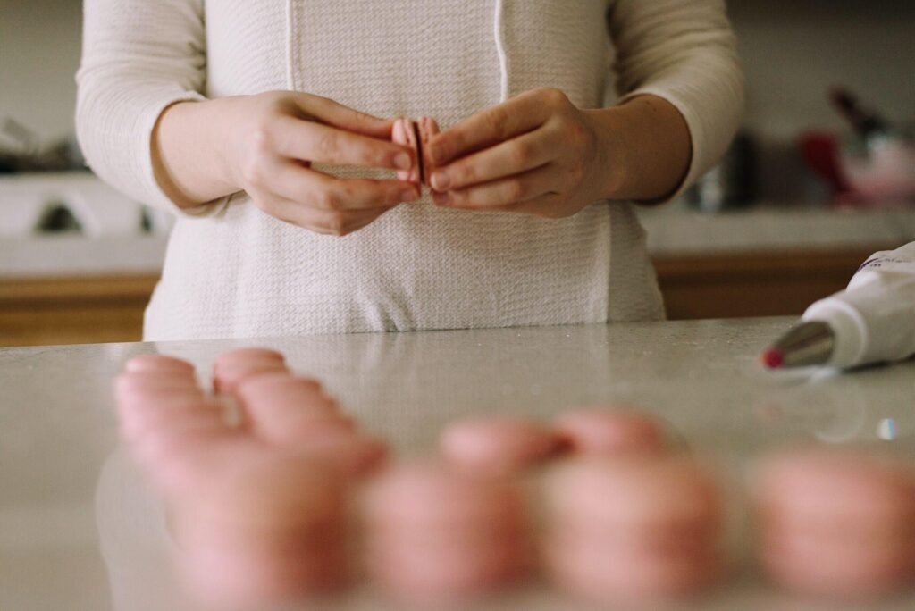 Person assembling pink macarons on a kitchen countertop, with a piping bag and additional macarons visible in the background, illustrating dessert preparation for French Buttercream Macarons.