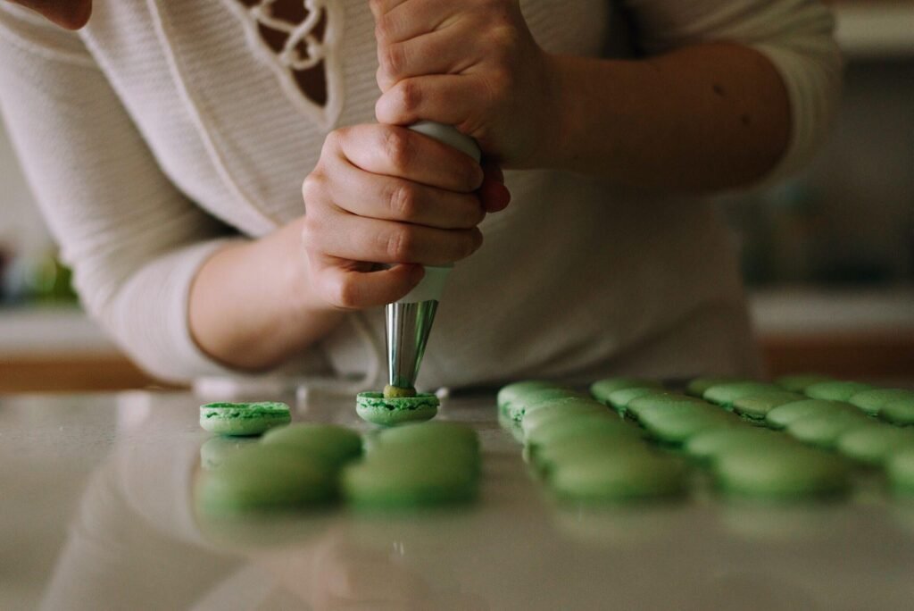 Person piping filling onto green macarons in a kitchen setting, highlighting the art of French Buttercream Macarons preparation.