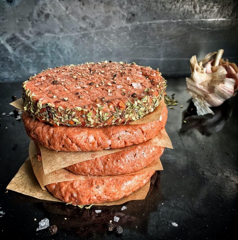 Stack of seasoned raw burger patties on parchment paper, with herbs and spices visible, against a dark countertop background, highlighting culinary preparation for a recipe.