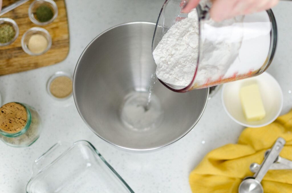 Hand pouring flour into a mixing bowl, surrounded by ingredients and utensils for baking chocolate chip cookies, including butter, measuring spoons, and spices on a countertop.