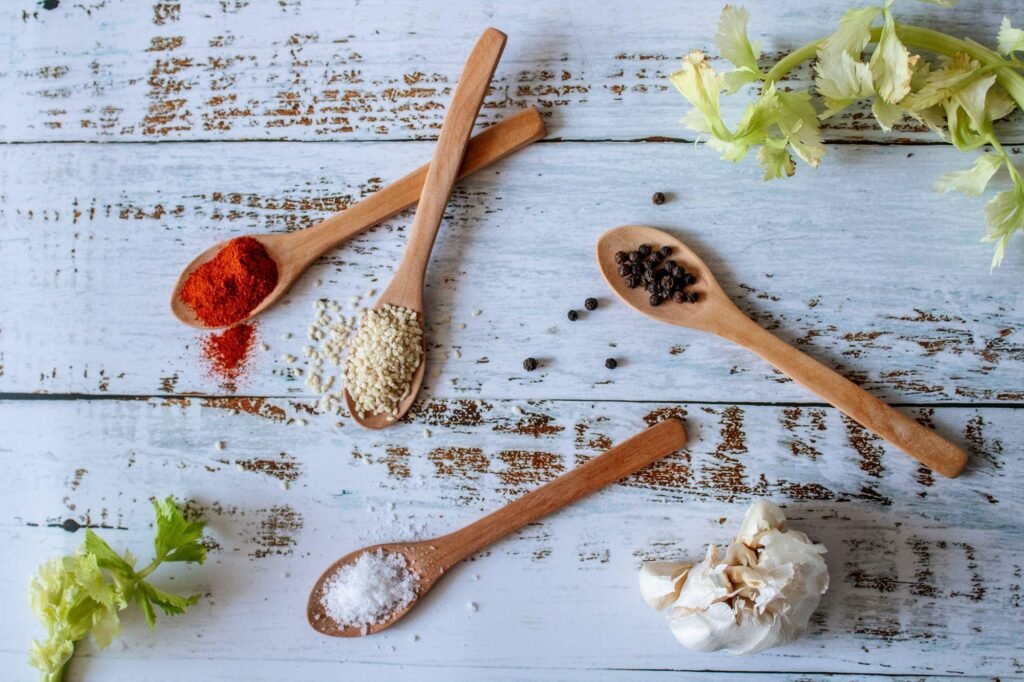 Wooden spoons with various spices including paprika, sesame seeds, black pepper, and salt, alongside fresh garlic and celery on a rustic wooden surface, illustrating ingredients for BBQ Chicken Pizza.