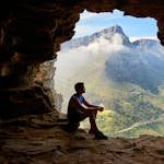 A man sitting in a cave overlooking a majestic mountain landscape under daylight.
